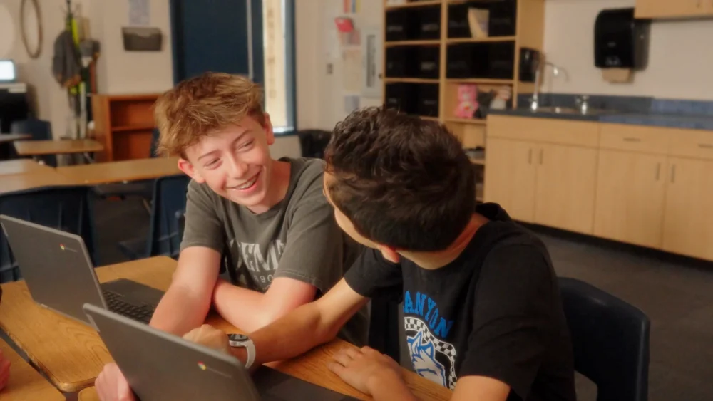 Two boys laughing together over a Chromebook at their desk.