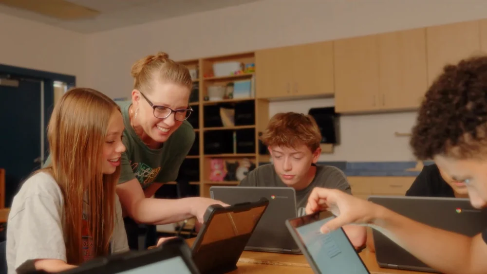 A teacher pointing at a student's tablet while four kids work around a classroom table.